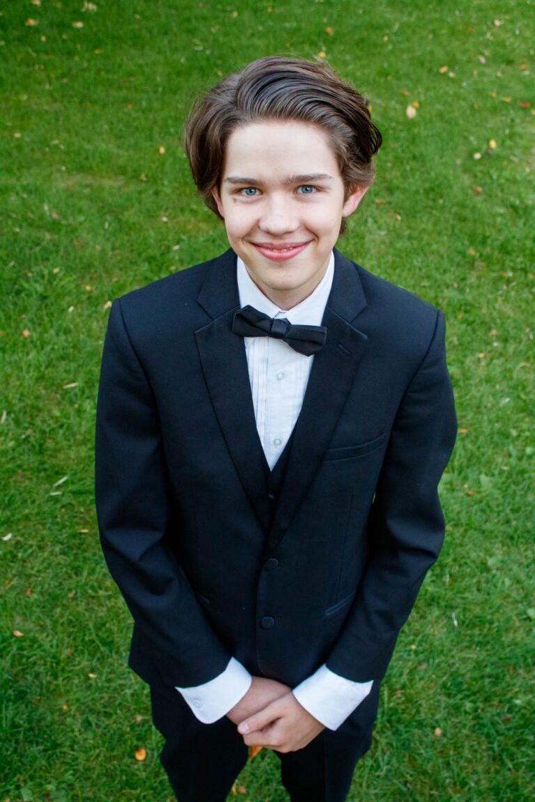 Boy in tuxedo smiling outdoors, dressed for a formal occasion or celebration.