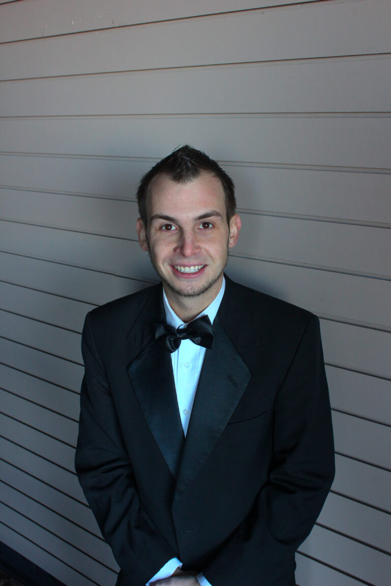 Man in formal tuxedo smiling at A & B Productions event, standing against a modern wall.