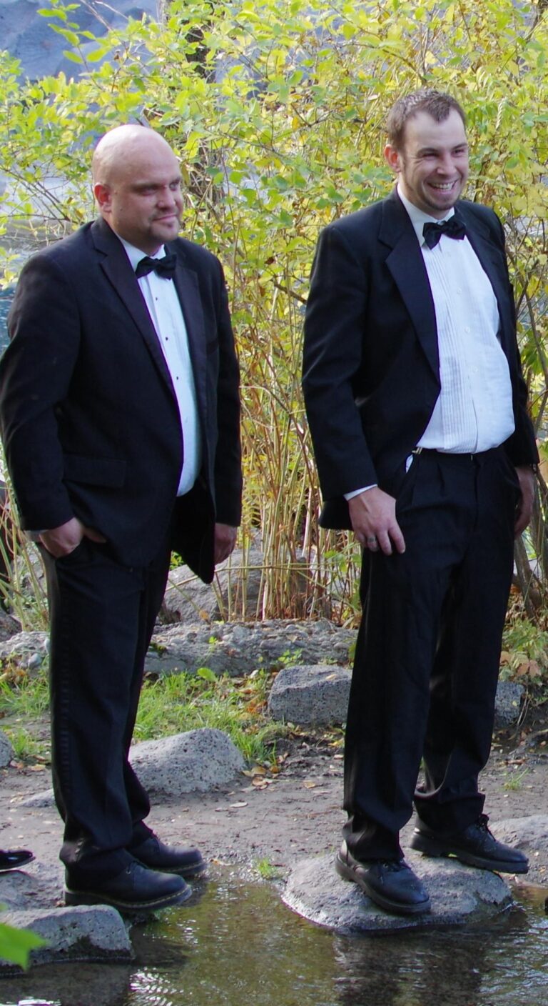 Men in formal tuxedos enjoying outdoor event near water and lush foliage.
