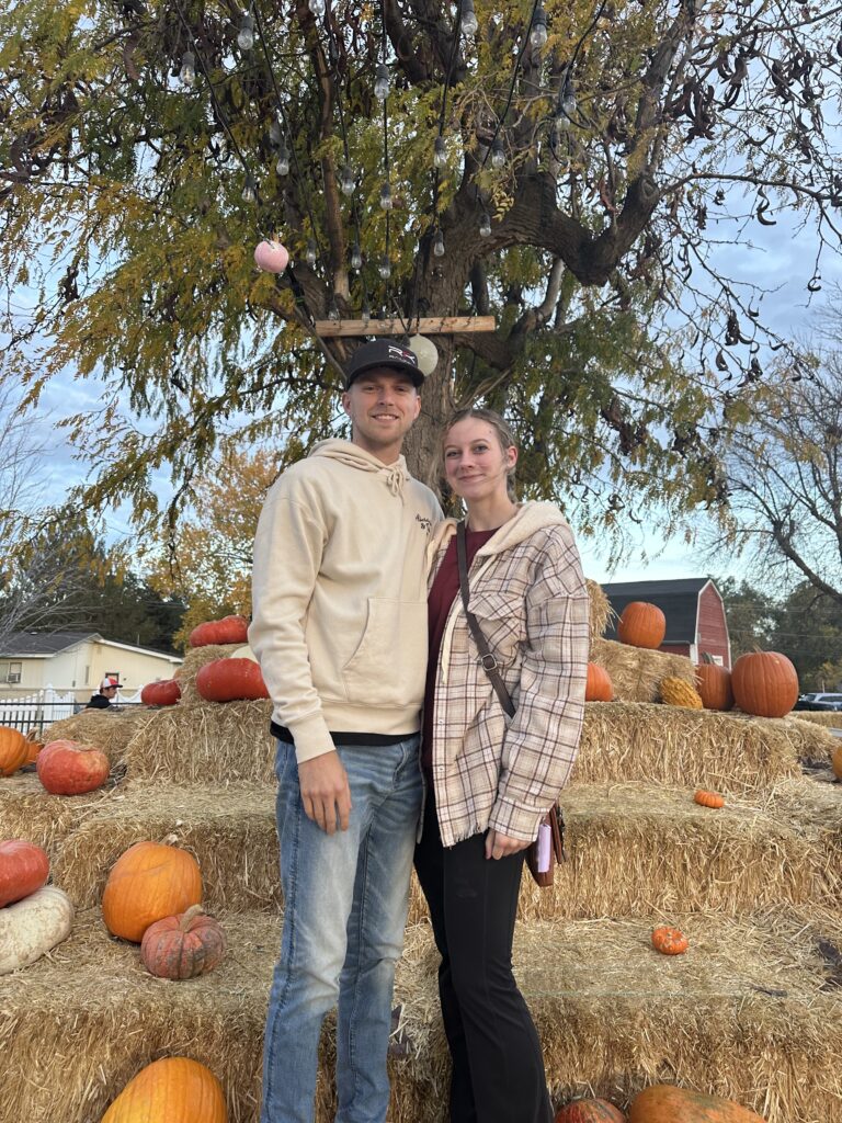 Couple standing in front of a fall pumpkin patch with hay bales and pumpkins, celebrating autumn