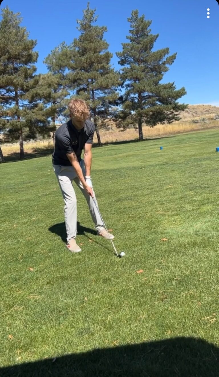 Person practicing golf swing on lush green course with trees in background.