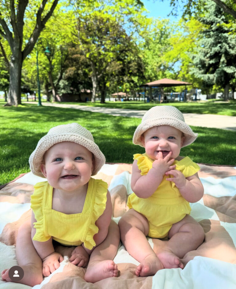 Twin babies in yellow outfits and hats sitting on a blanket outdoors in a park.