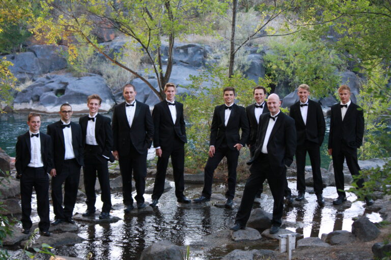 Group of men in tuxedos standing in a river with trees and rocks in the background.