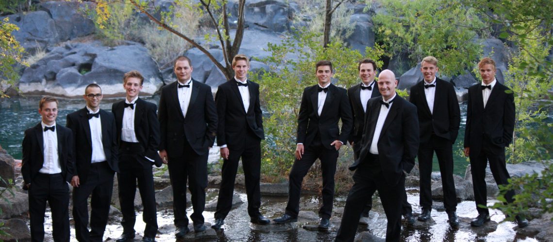 Group of men in tuxedos standing in a river with trees and rocks in the background.